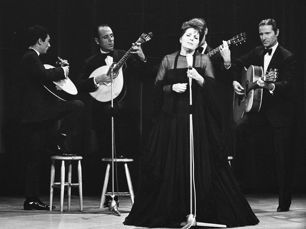 Amália Rodrigues w długiej czarnej sukni śpiewająca do mikrofonu na scenie, a wokół niej siedzą gitarzyści w ciemnych garniturach.
