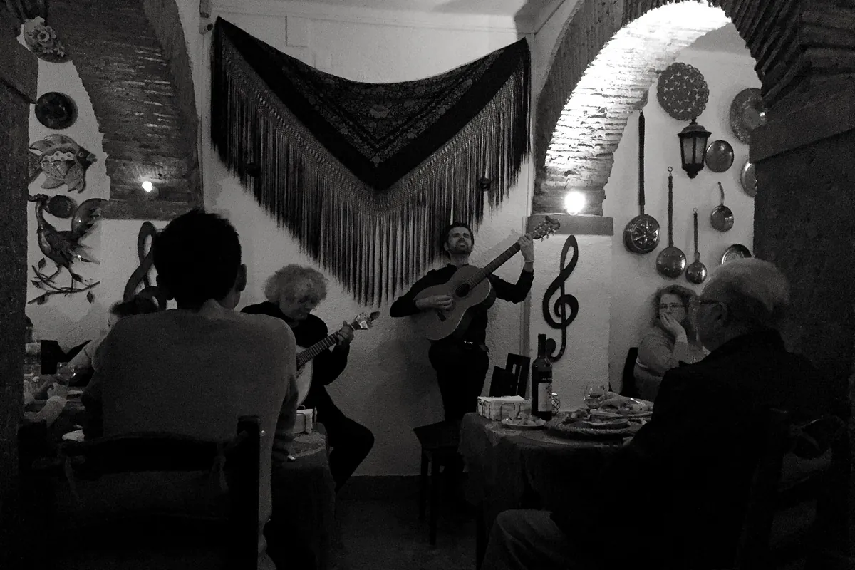 Przygaszona, łukowa sala w Lizbonie z czarnym szalem z frędzlami na ścianie i dwoma gitarzystami grającymi wśród gości przy małych stolikach.