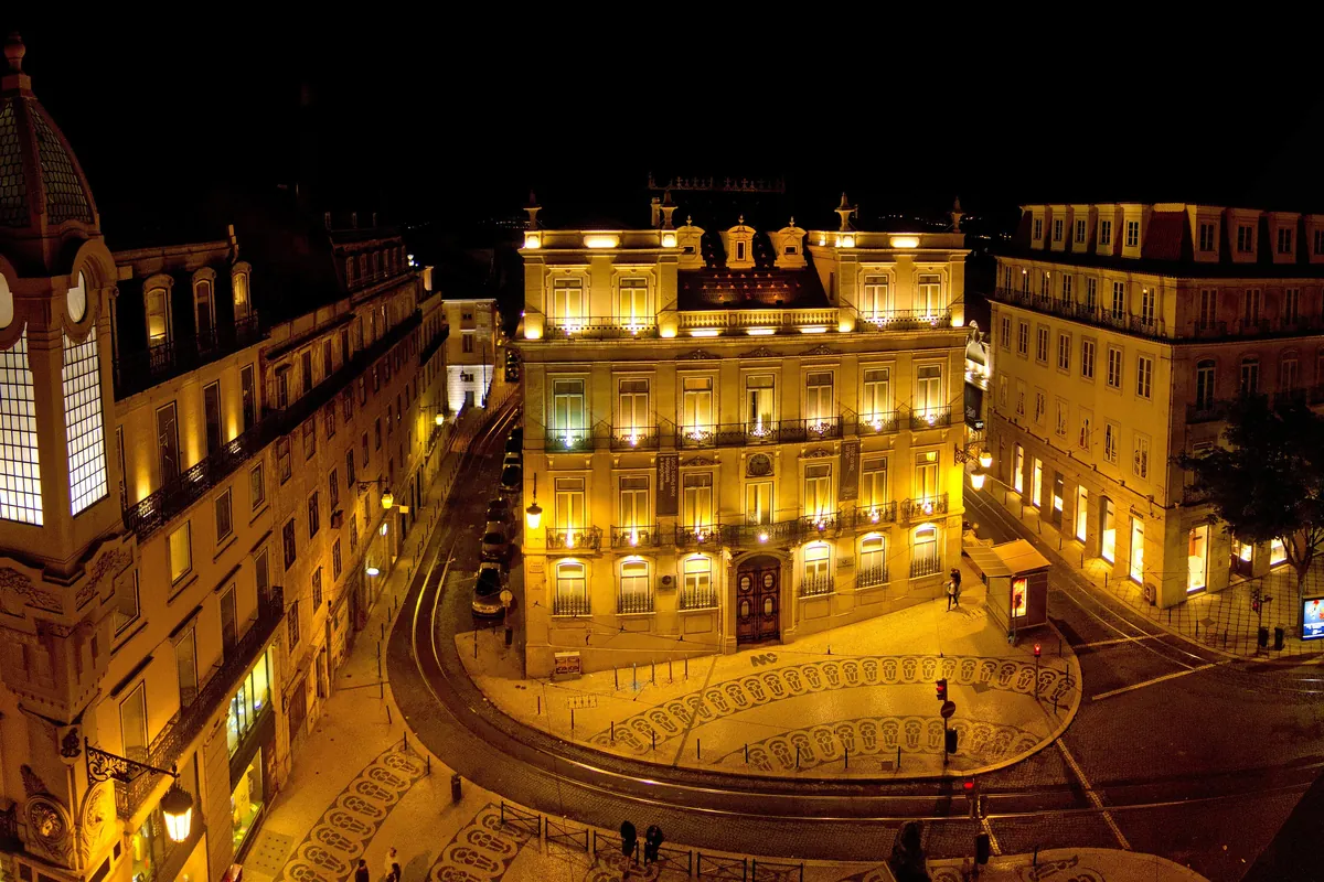 Plac Largo do Chiado nocą, z podświetlonymi żółtymi fasadami i wieżą zegarową po lewej stronie.