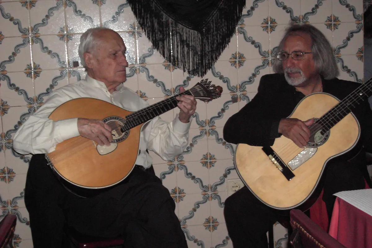 Portugalska gitara i gitara akustyczna grają obok siebie przed ścianą z kafli, na której wisi czarny szal z frędzlami.
