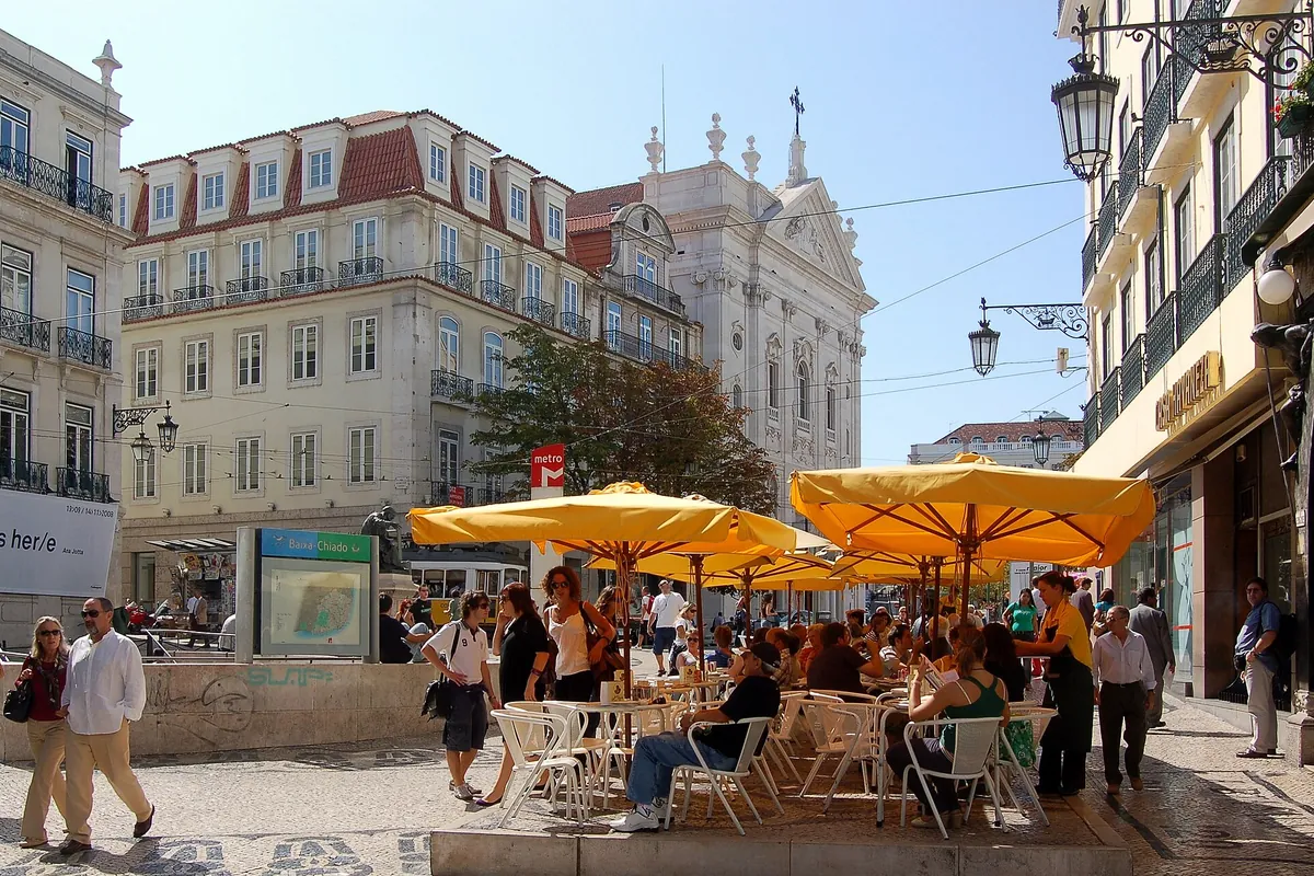 Żółte parasole nad stolikami kawiarnianymi na Largo do Chiado, a za nimi blade zabytkowe budynki.